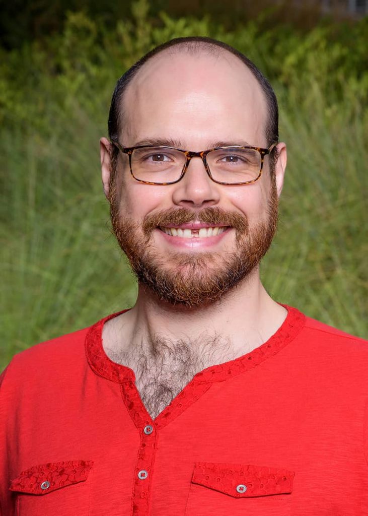 Portrait photograph of a balding person with glasses and a beard wearing a red, feminine-coded top.